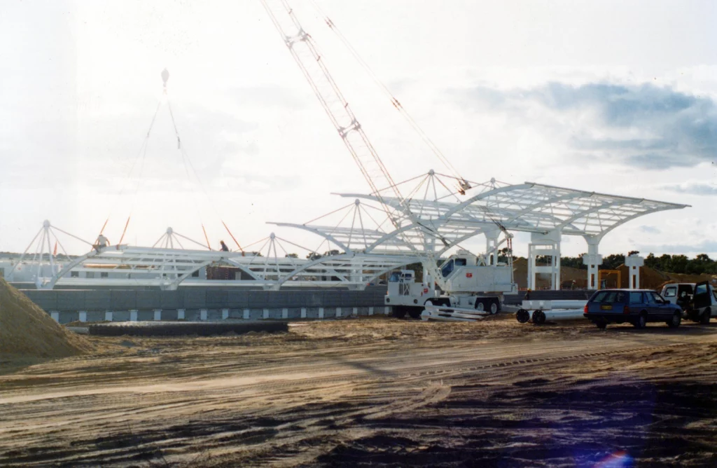 A large white metal structure is being assembled at a construction site with cranes and vehicles. The ground is sandy, and the sky is partly cloudy. Several workers and equipment are visible around the site.