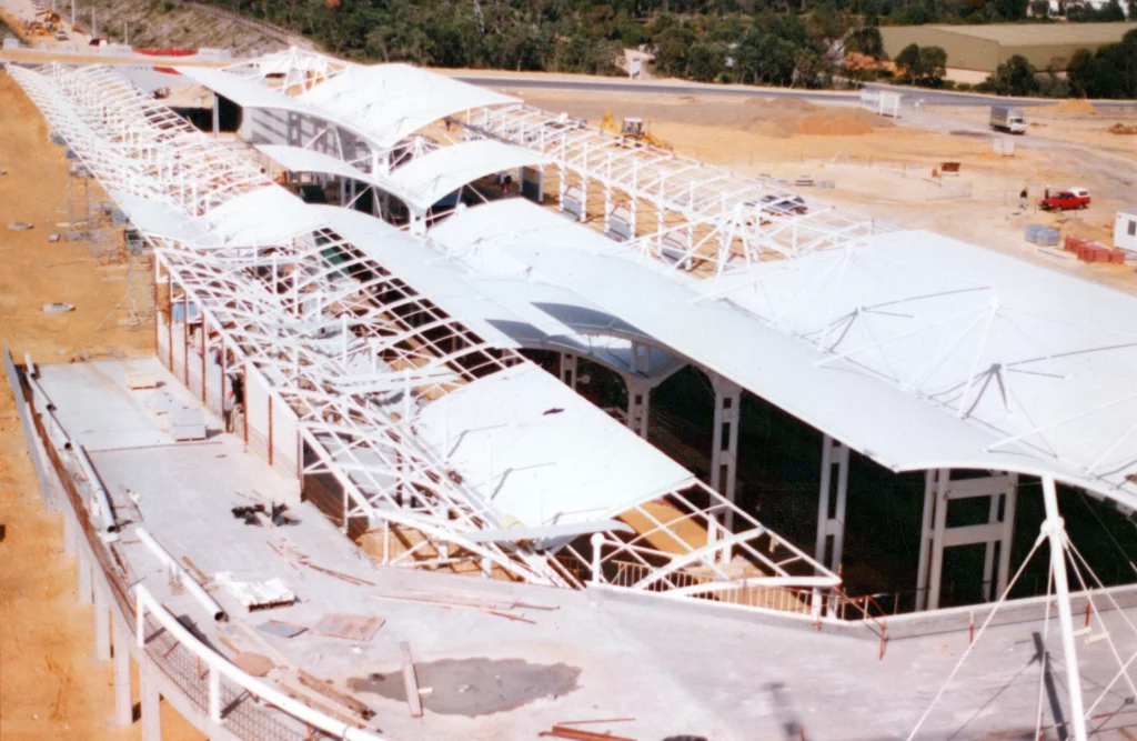 Aerial view of a large building under construction with a white, wavy steel and fabric roof structure partially installed over a sandy construction site. Scaffolding and construction materials are visible.