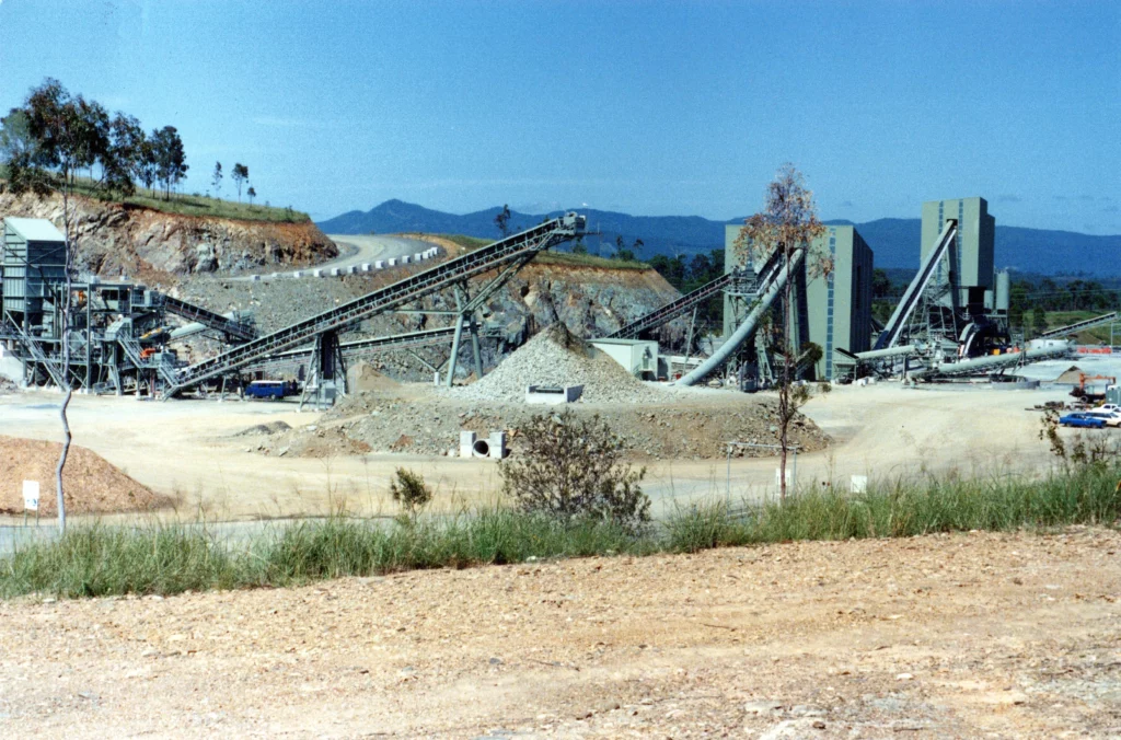 A large industrial quarry site with conveyor belts, machinery, and piles of gravel, set against a backdrop of trees and distant mountains under a clear blue sky.