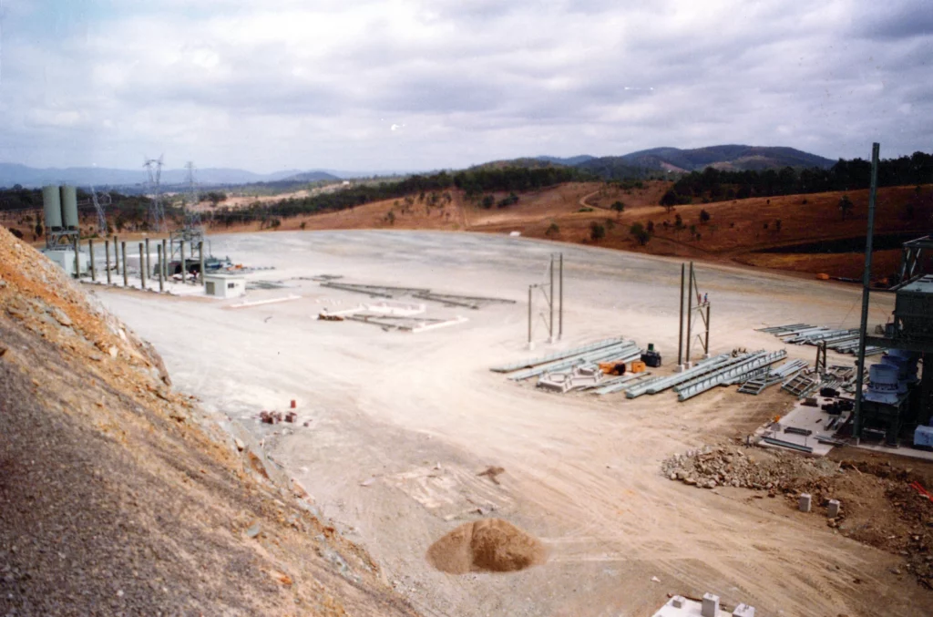 A large, mostly barren construction site with scattered industrial equipment and metal pipes, surrounded by dry, hilly terrain under a cloudy sky. Sparse vegetation and distant hills are visible in the background.