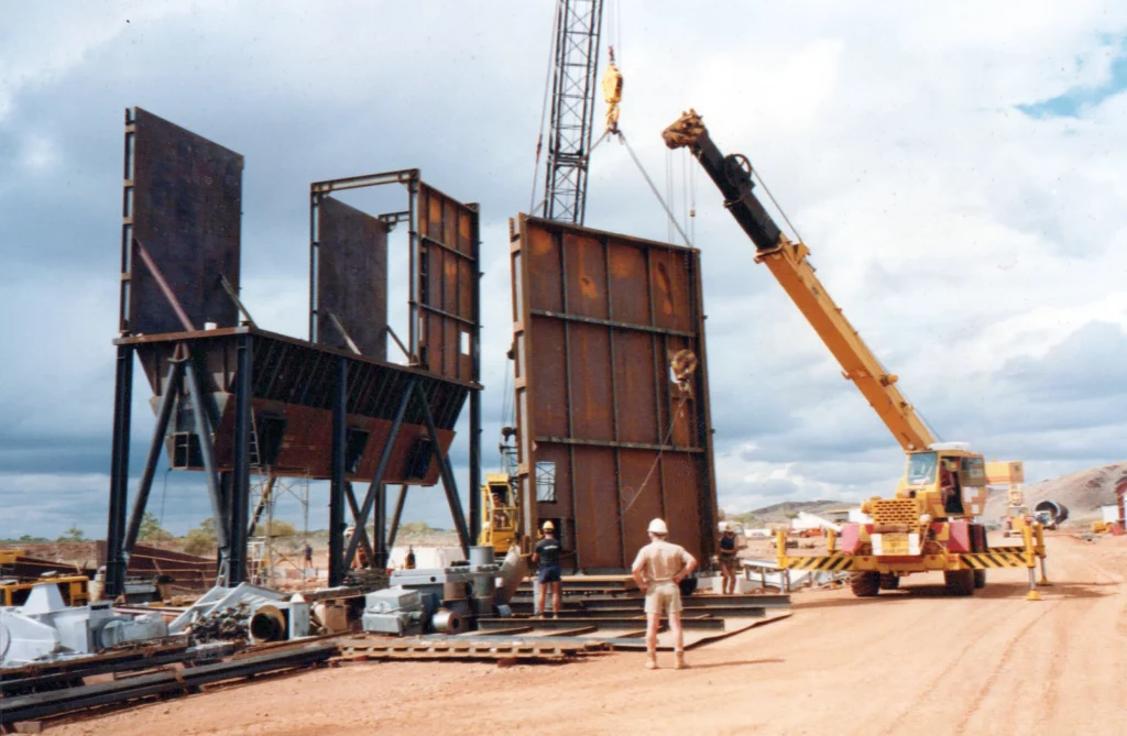 A crane lifts a large metal panel at a construction site with workers nearby, assembling tall steel structures on a dirt ground under a cloudy sky.