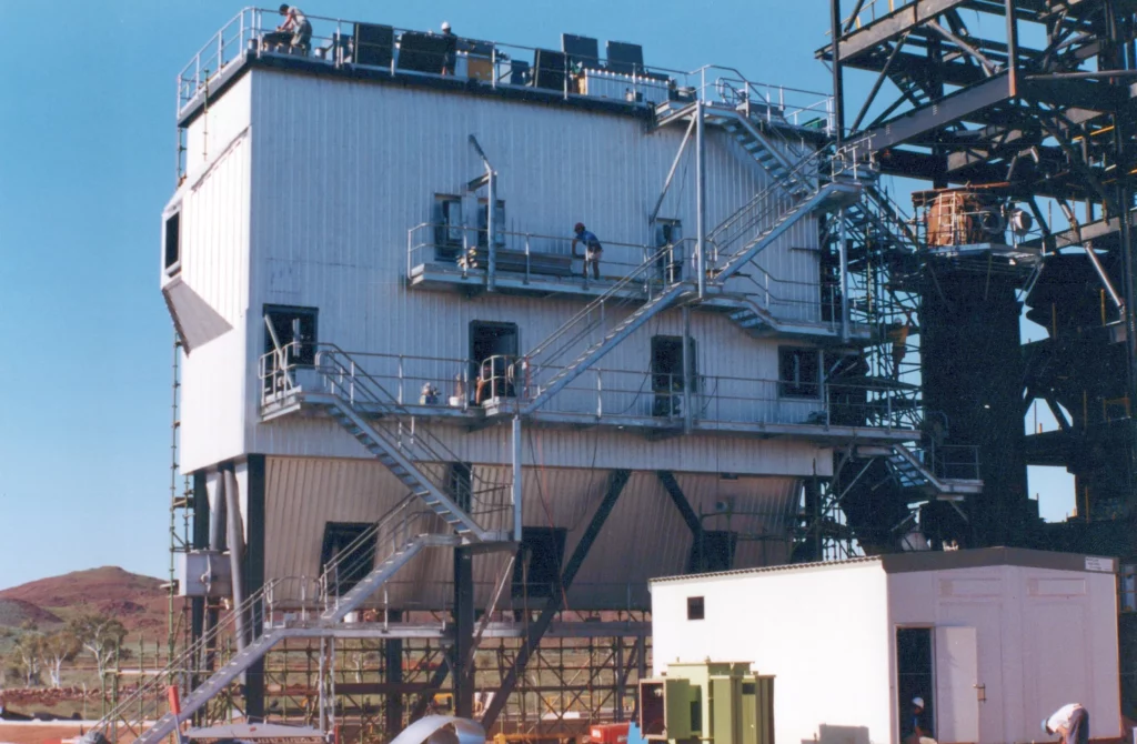 A large industrial building structure with exterior staircases and railings, elevated and supported by metal beams, stands against a clear blue sky. People are visible on the stairs and ground below.