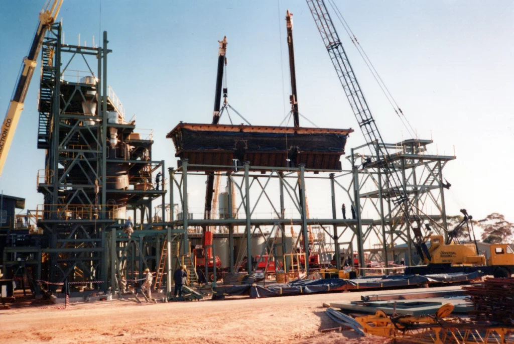 A large industrial construction site with cranes lifting a heavy structure onto a steel framework. Workers and machinery are visible on the sandy ground, with scattered construction materials in the foreground.
