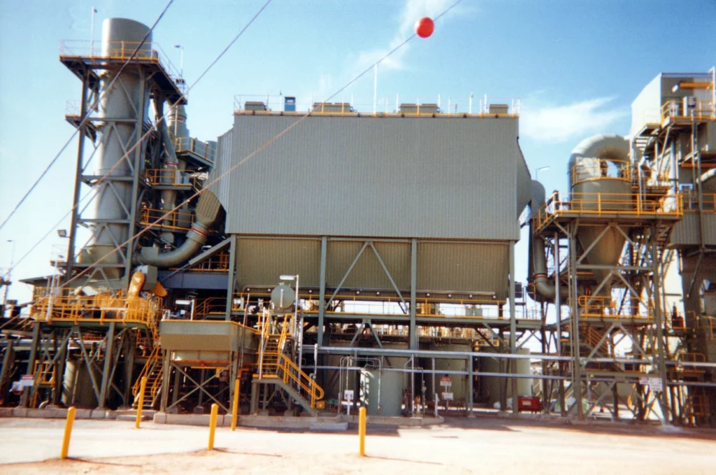 A large industrial facility with metal structures, vertical tanks, pipes, spiral staircases, and yellow safety railings under a clear blue sky. A red ball marker hangs from overhead wires in the foreground.