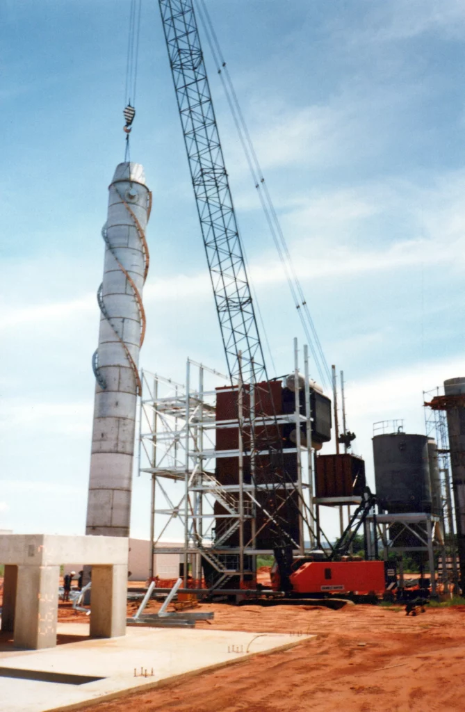 A crane lifts a large, spiral-shaped metal structure at a construction site with steel frameworks, tanks, and equipment visible in the background under a blue sky.