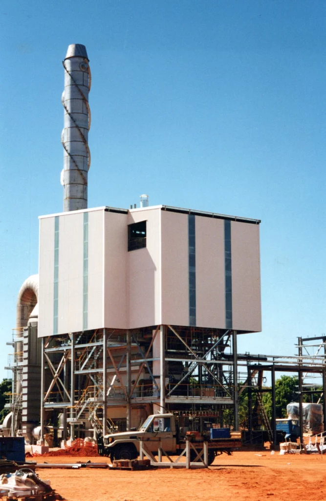 A large industrial facility with a tall, spiral-wrapped smokestack stands on a red dirt site under a clear blue sky. Metal structures, pipes, and a truck are visible around the building.
