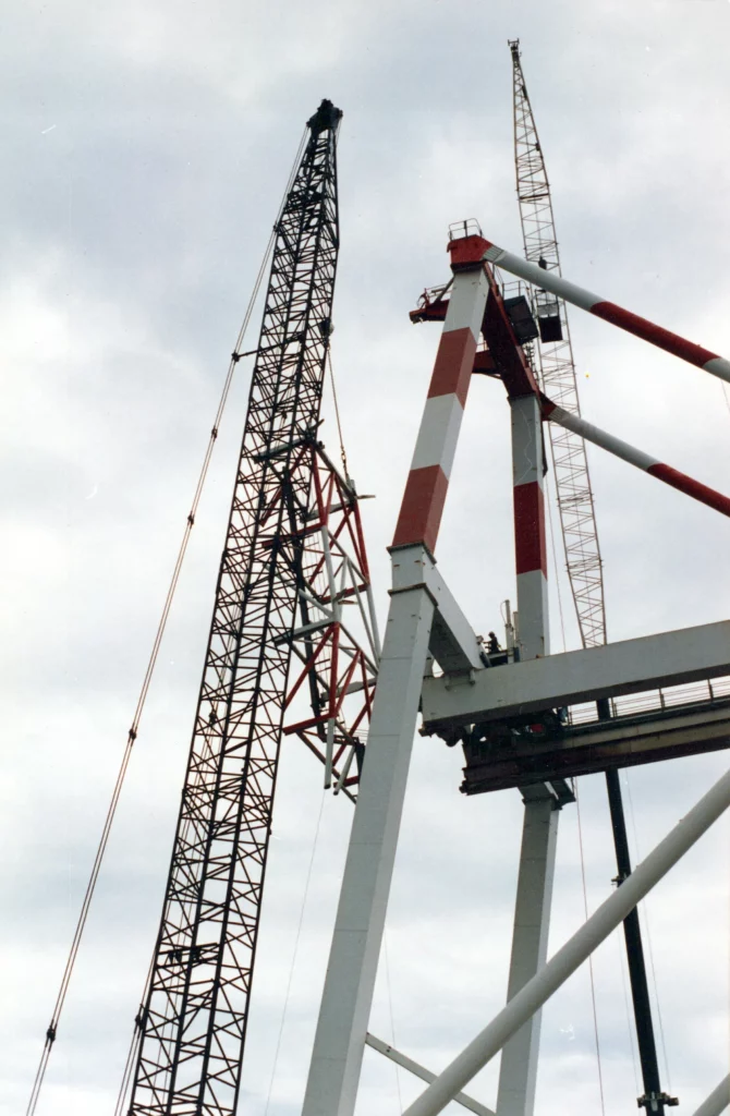 A close-up view of large construction cranes, including a black lattice crane and a red-and-white crane, working against a cloudy sky. The cranes are lifting or assembling large metal structures.