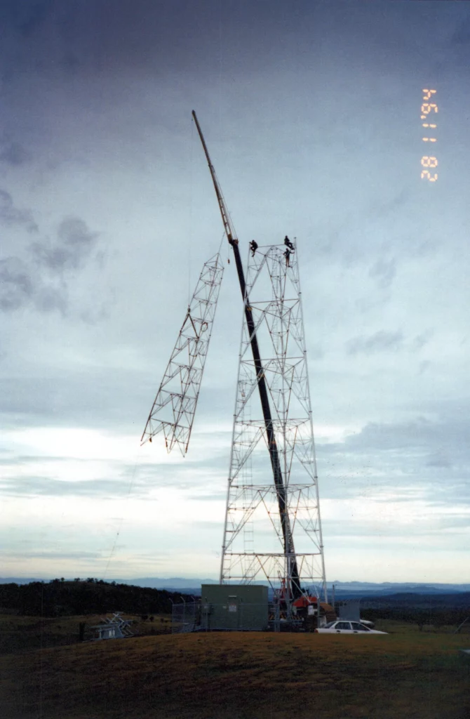 A tall radio tower under construction, with workers on top and a crane lifting a large metal section into place, set against a cloudy evening sky. The date "28. 1. 1994" is visible on the top right.