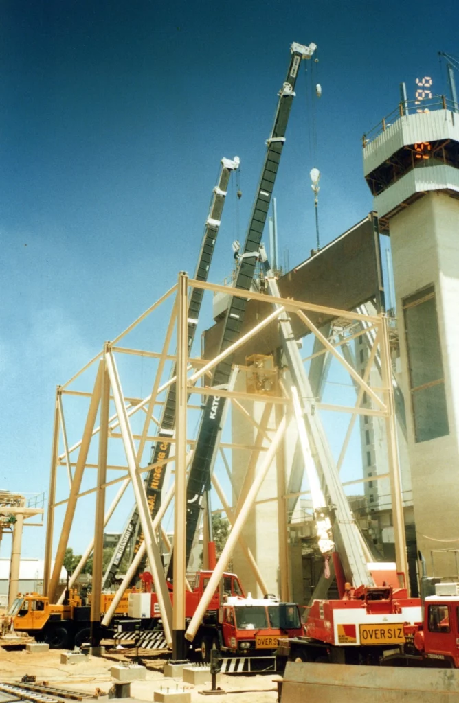 Construction site with multiple cranes lifting large steel beams. Workers are seen on elevated platforms, and various construction vehicles labeled "OVERSIZE" are positioned below. A partially built structure is in the foreground.
