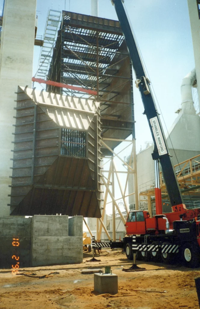 A large crane lifts a heavy, industrial metal structure at a construction site with steel frames, concrete, and scaffolding under a clear sky. The ground is covered in dirt and construction materials.