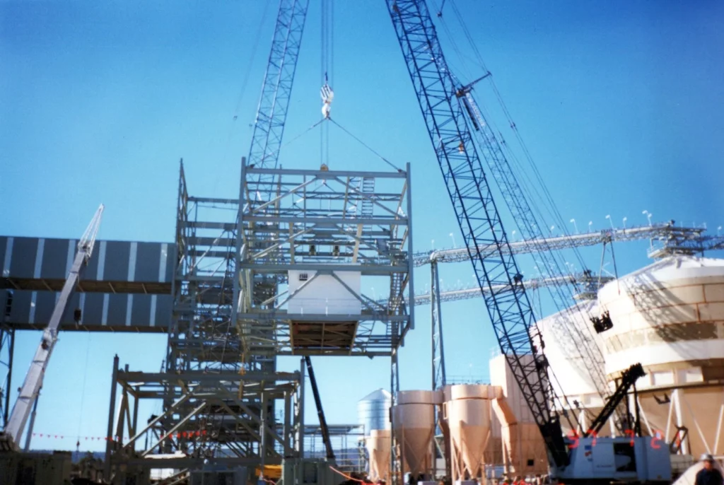 A large crane lifts a metal structure at an industrial construction site with steel frameworks, cylindrical storage tanks, and blue sky in the background.