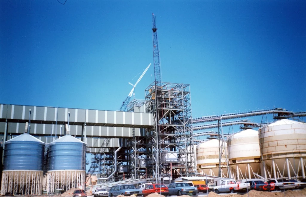 A large industrial facility under construction with scaffolding, cranes, and steel framework. Several cylindrical tanks and silos are visible, along with parked cars in the foreground under a clear blue sky.