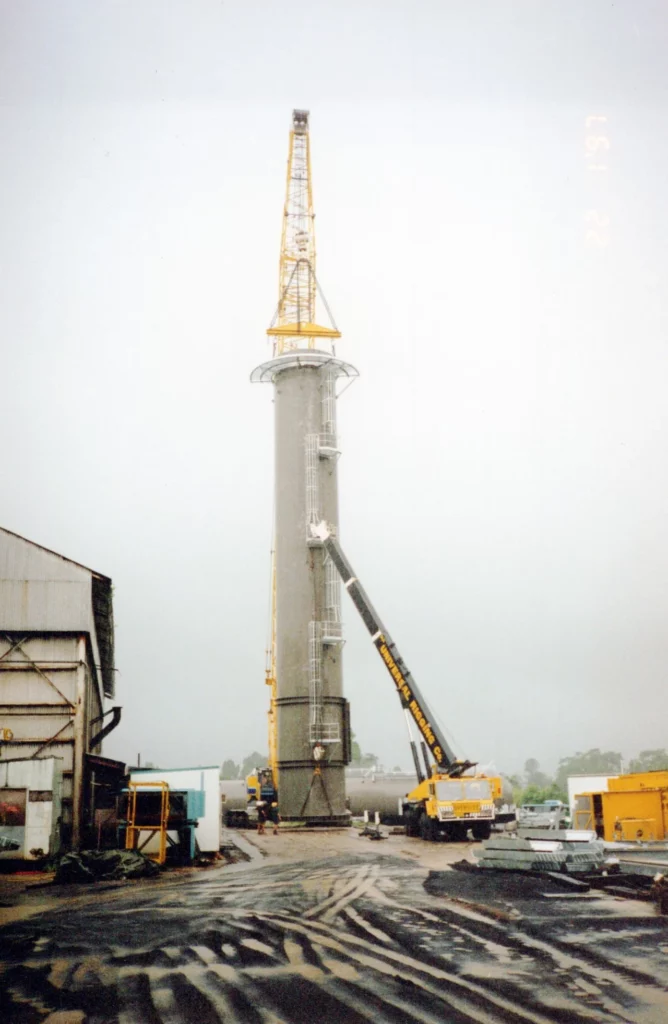 A tall industrial tower is being installed by two large yellow cranes, with workers and equipment at the base. The scene takes place at an outdoor construction site near warehouse buildings.