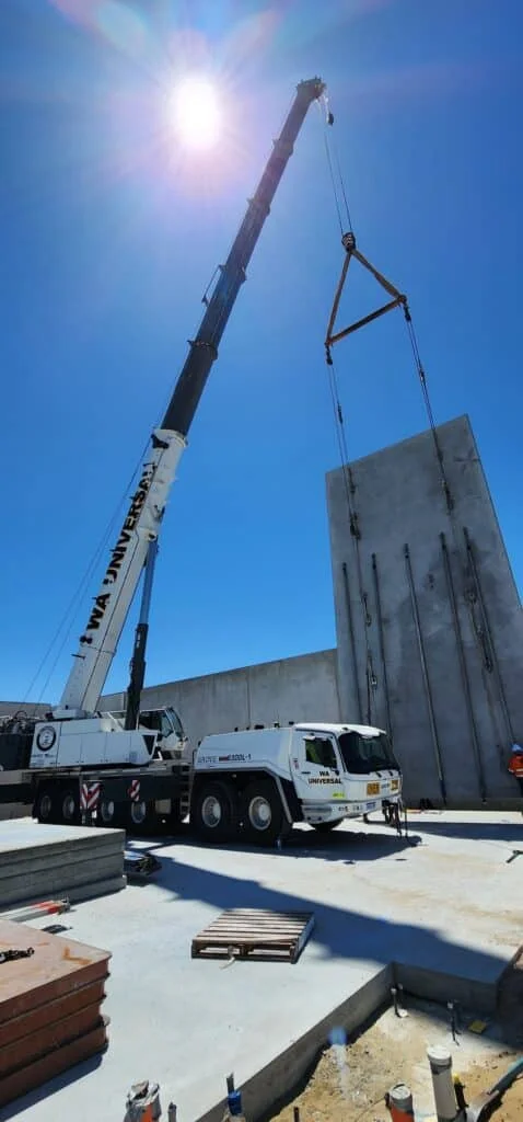 A large crane lifts a tall concrete wall panel at a construction site under a bright blue sky with the sun shining overhead. Construction materials and shadows are visible on the ground.