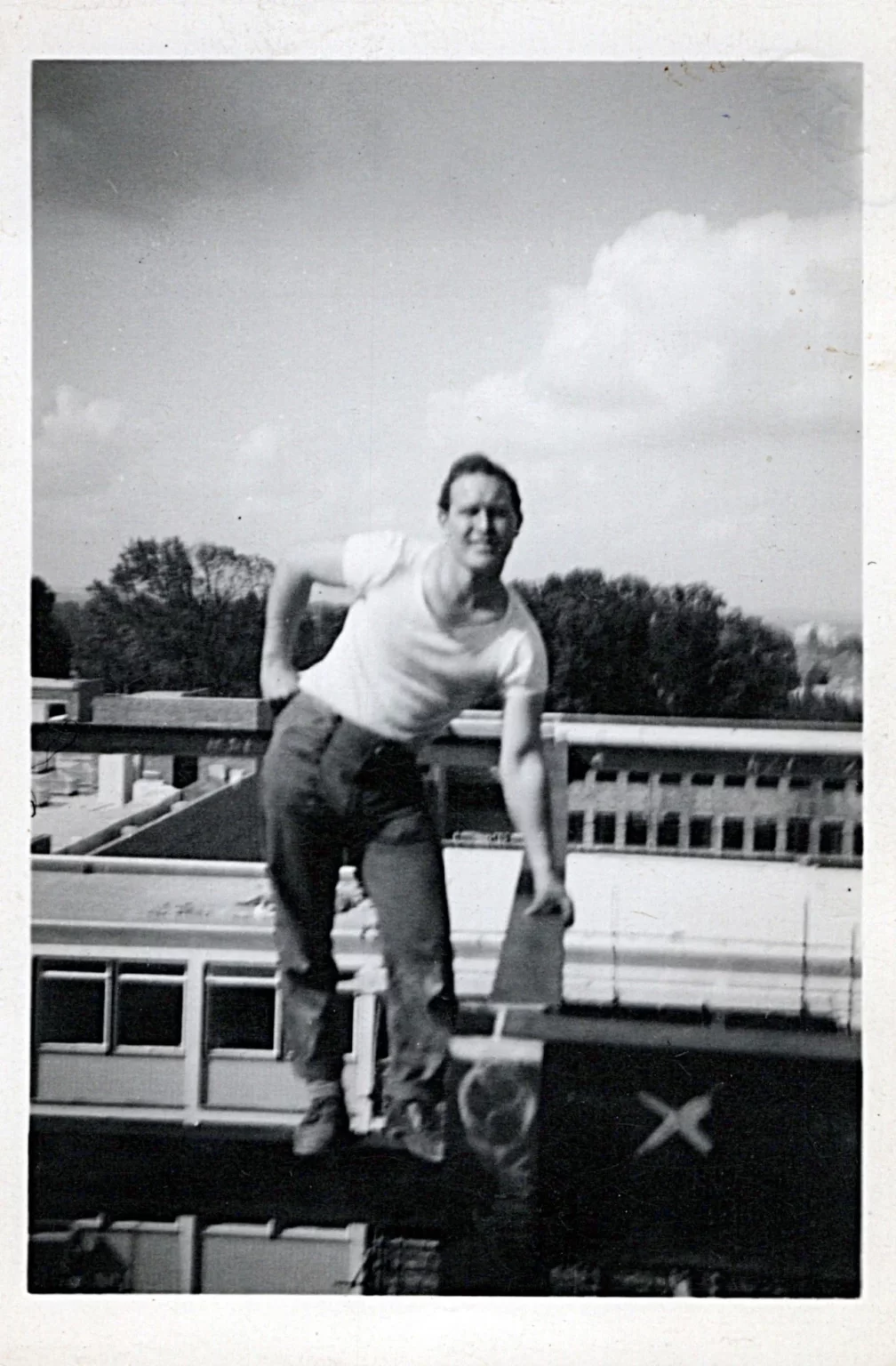 A person in a white t-shirt and jeans balances on a railing or beam above a building, with trees and a partly cloudy sky in the background. The image is in black and white and has a vintage feel.