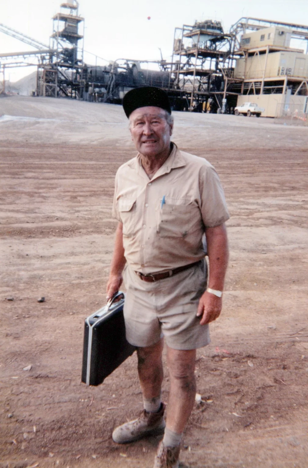 An older man in a tan work outfit and black cap stands on a dirt lot, holding a briefcase. His legs and boots are dusty. Industrial structures and equipment are visible in the background.
