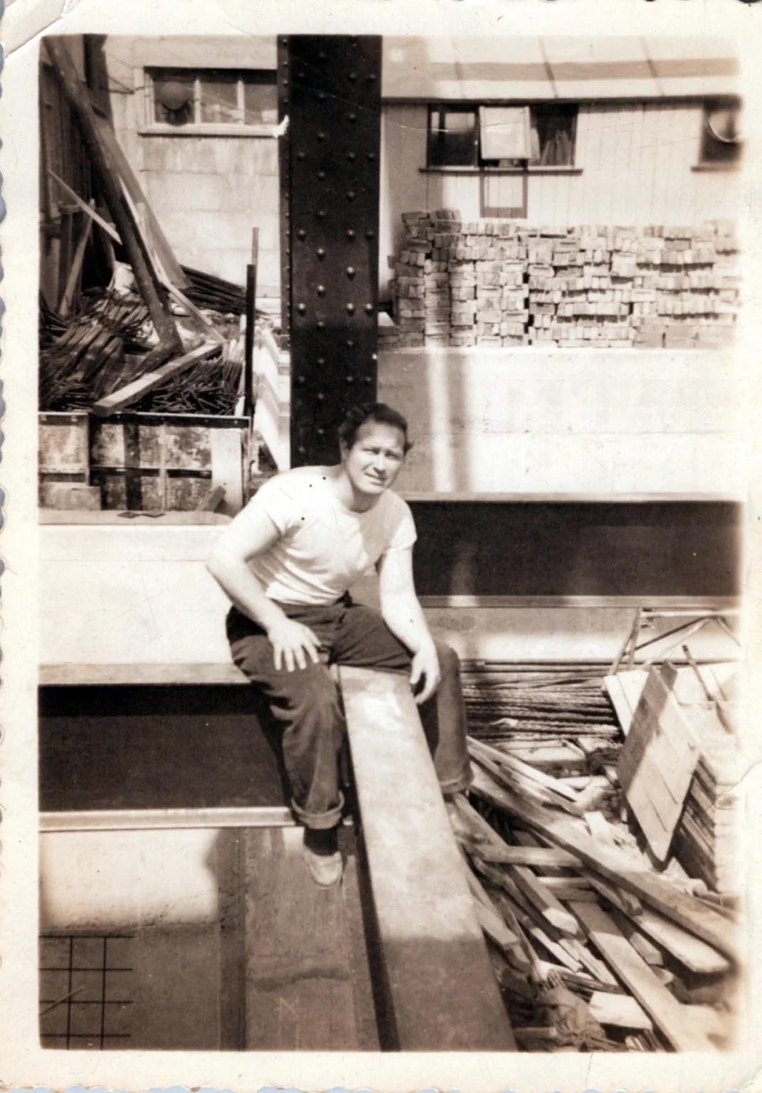 A person in a white T-shirt and rolled-up jeans sits on a steel beam at a construction site, surrounded by building materials and stacks of bricks. The photo is in black and white, creating a vintage look.