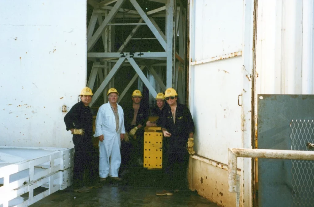 Five workers wearing hard hats and coveralls stand together near a large industrial door, with one person in white coveralls and the others in dark outfits. They are posing around a yellow metal object.