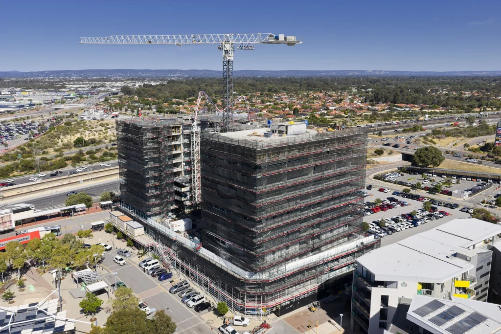 A tall building under construction, covered in scaffolding, with a large crane on top. Surrounding the site are roads, parked cars, and suburban housing in the distance under a clear blue sky.