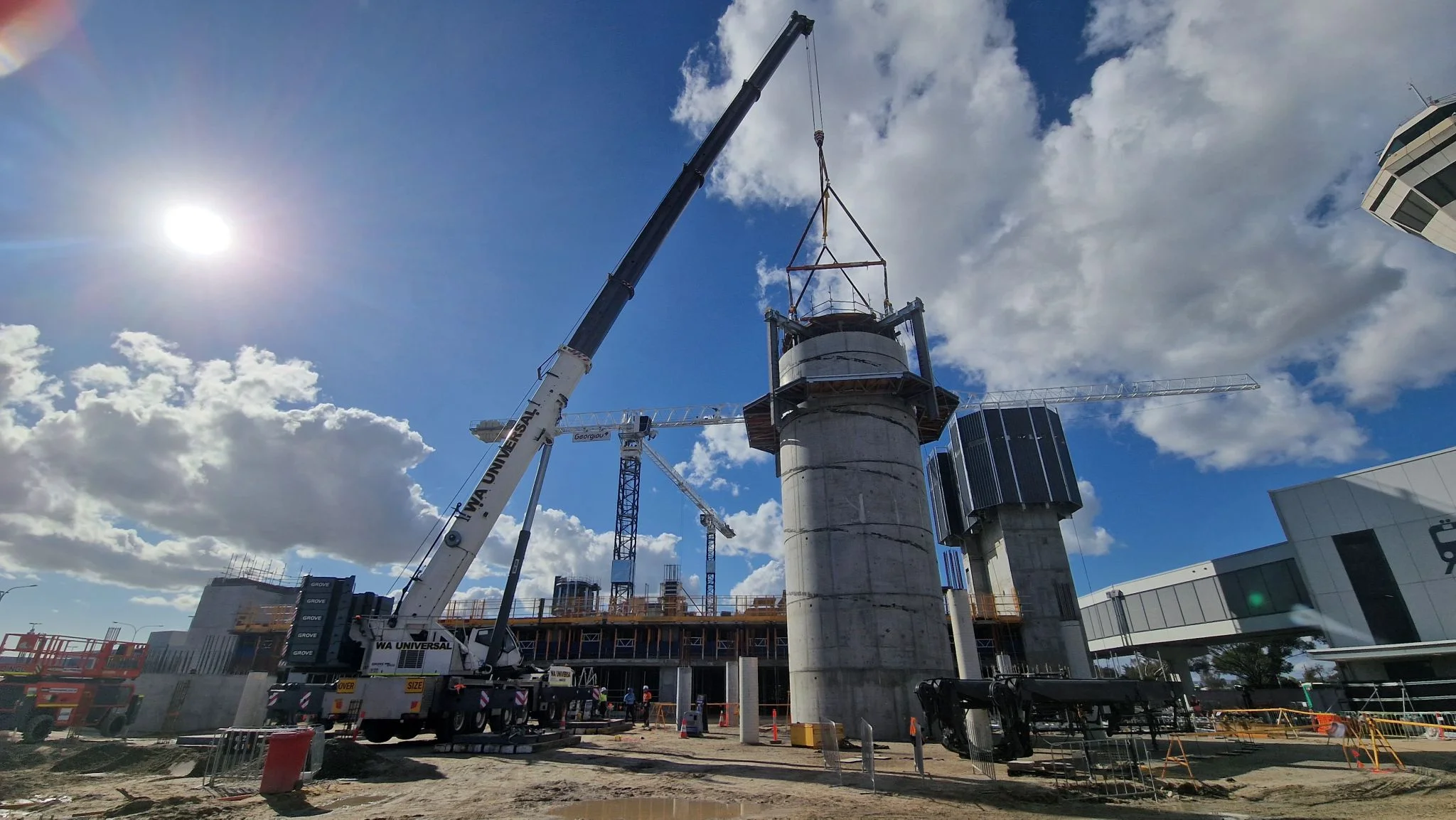 A construction site with tall concrete structures, cranes lifting materials, and workers present. The sun is shining, with clouds in the blue sky and a partially built building in the background.