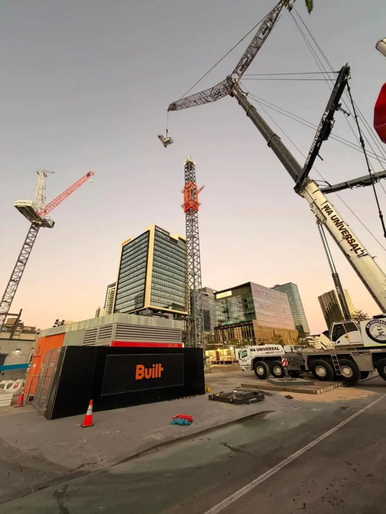 Construction site with cranes and modern glass buildings at sunset. A large white crane truck and portable building labeled "Built" are in the foreground. Traffic cones and scattered equipment are visible on the ground.