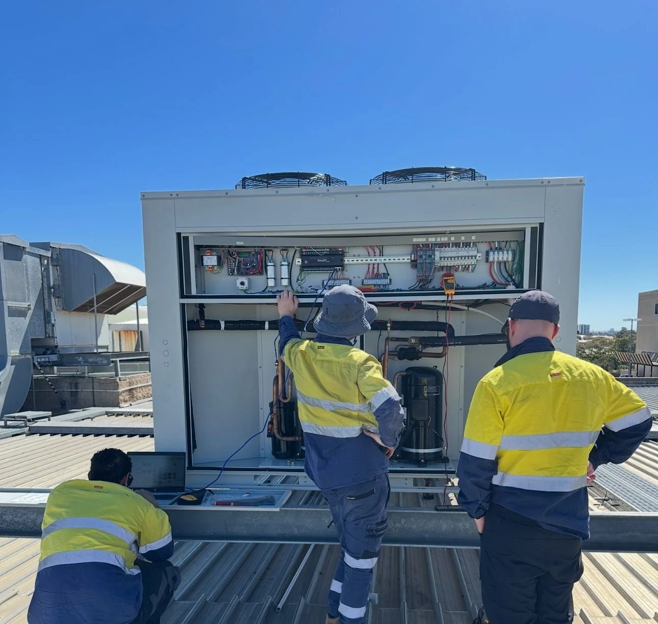 Three workers in yellow and navy safety uniforms inspect and work on the controls of a large industrial HVAC unit on a rooftop under a clear blue sky.