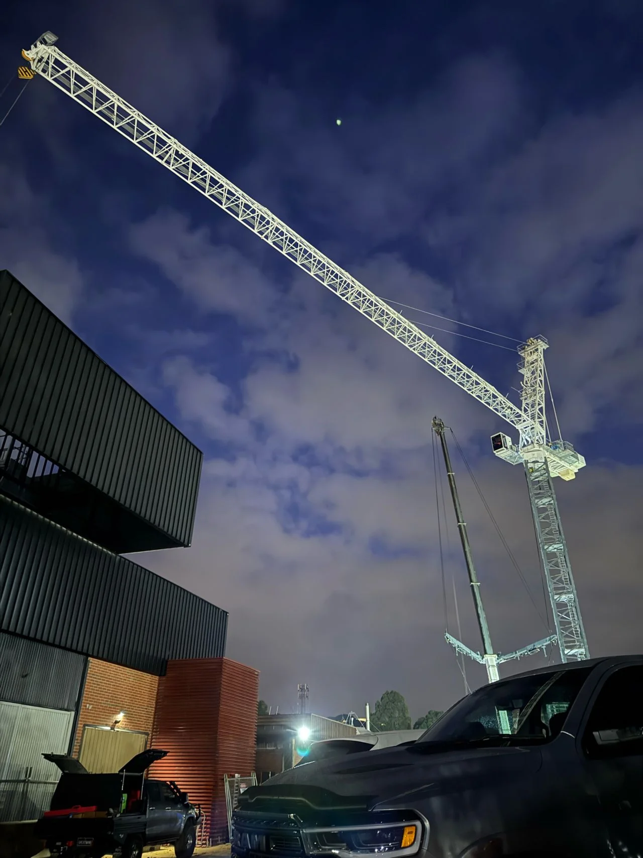 A tall construction crane illuminated at night stands over parked vehicles and modern buildings under a cloudy, blue-tinged sky.