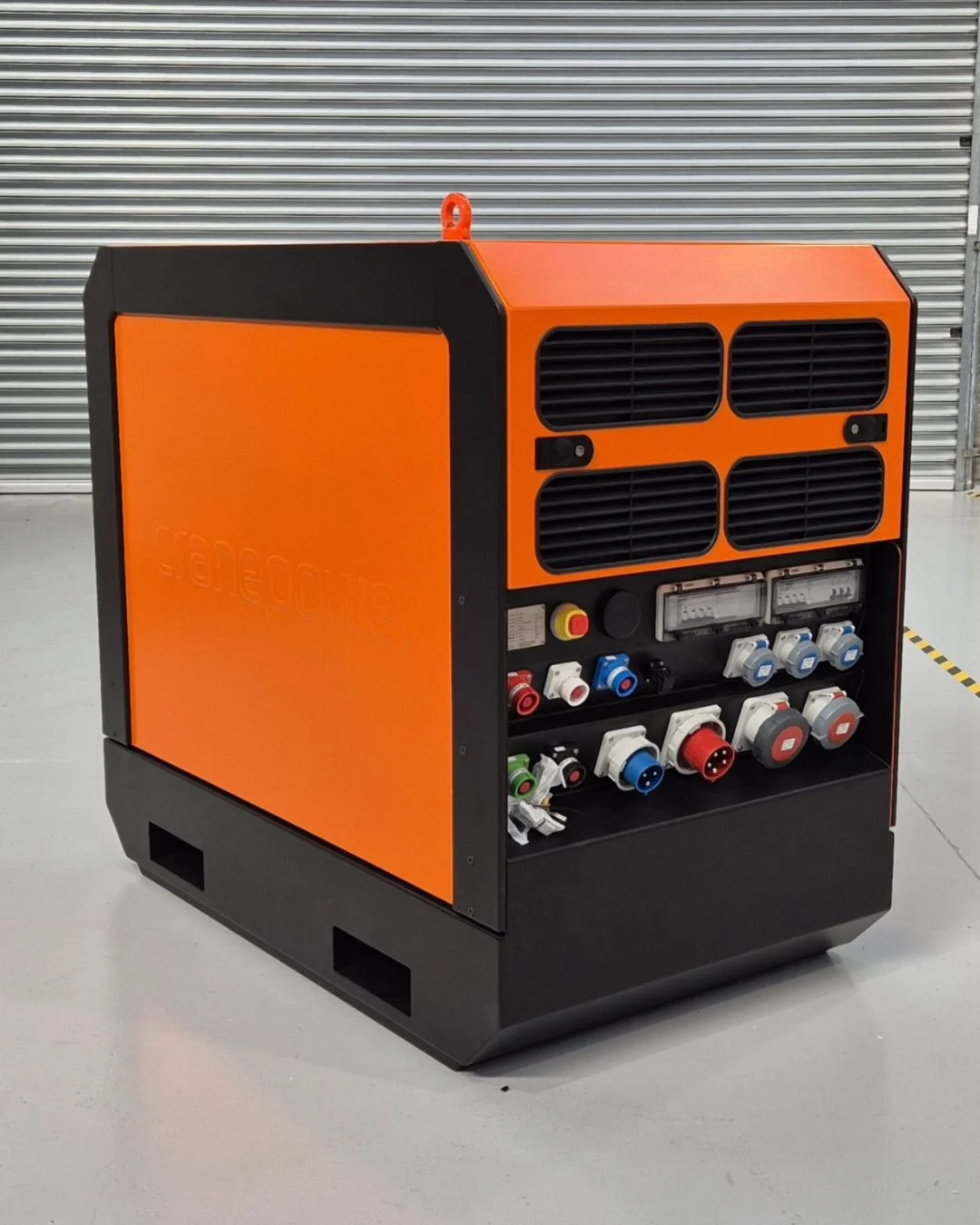 Large industrial orange and black power distribution unit with multiple electrical sockets, switches, and cooling vents, placed on a smooth indoor floor in front of a gray corrugated metal wall.