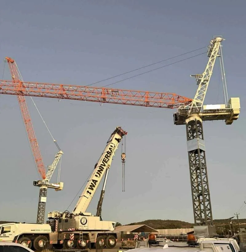 A mobile crane labeled "IWA UNIVERSAL" lifts part of a tall construction crane, while another orange tower crane is seen in the background against a clear sky at a construction site.