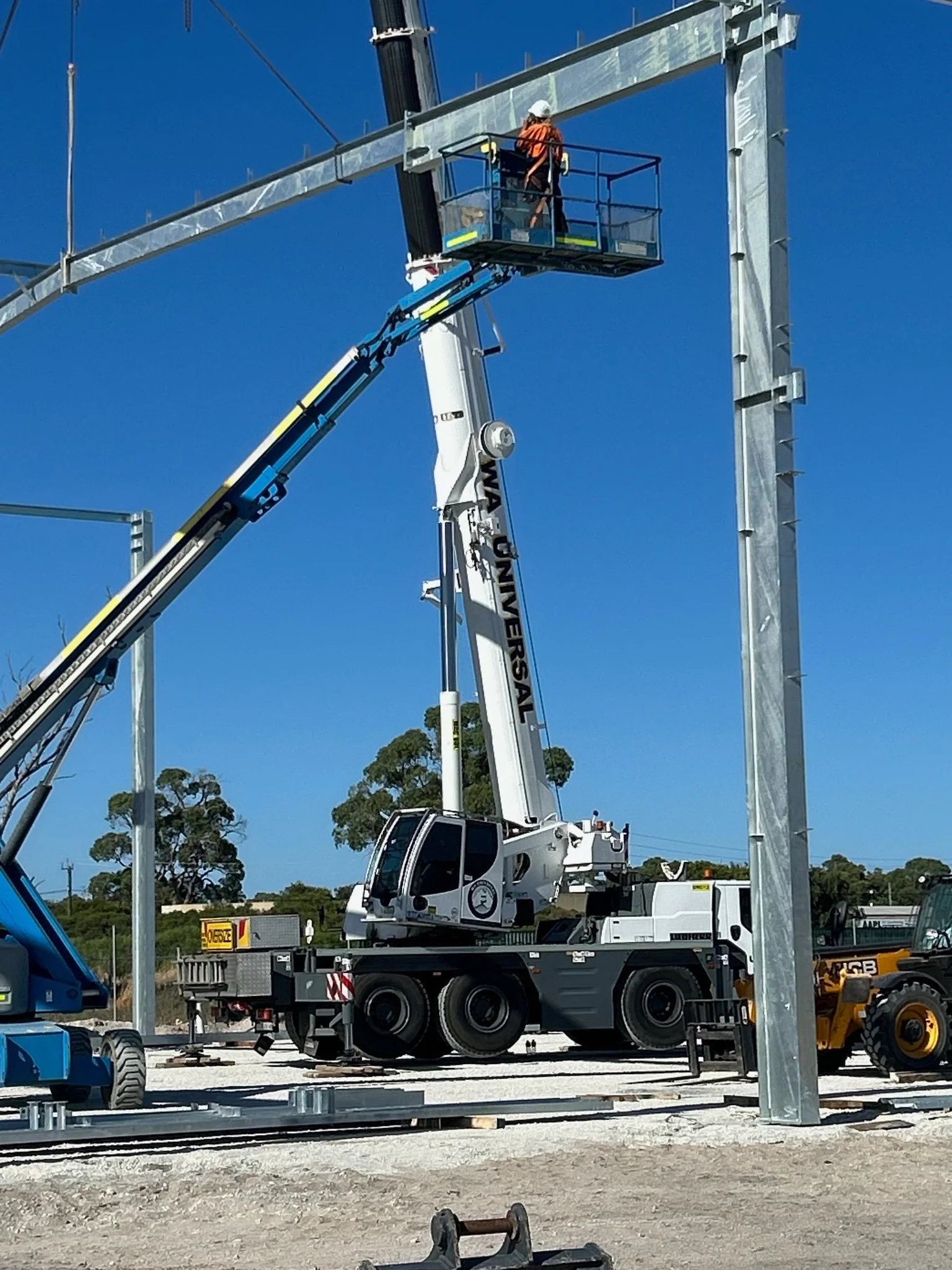 A construction worker in a safety harness stands on a raised platform, working on a steel frame structure, with a large crane and other machinery on site under a clear blue sky.