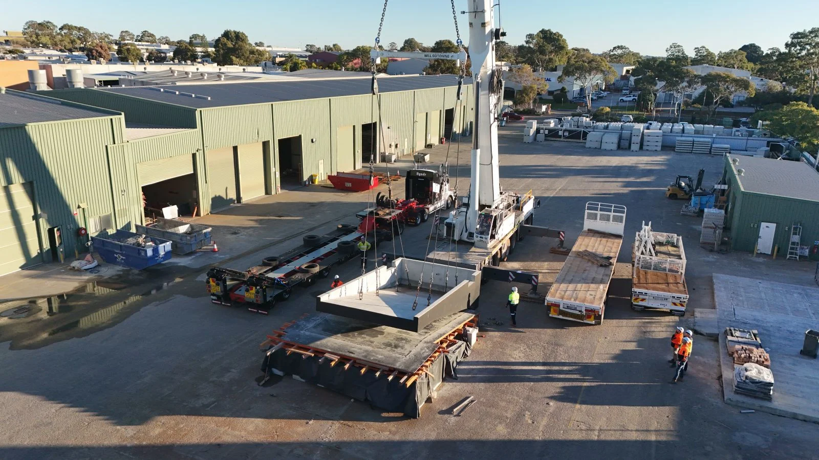 A crane lifts a large concrete panel at an industrial facility with green warehouses. Several workers in safety gear guide the operation, and trucks with building materials are parked nearby.