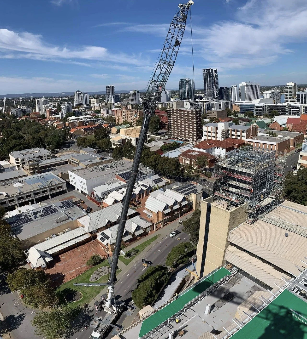 A tall crane lifts materials to the rooftop of a high-rise building under construction in an urban area, with other city buildings, roads, and scattered trees visible below and in the background.