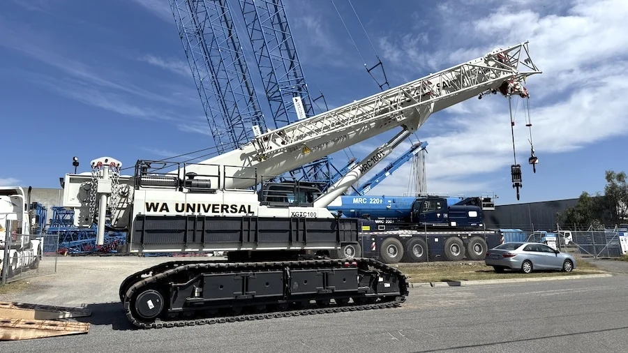 A large white crane labeled "WA UNIVERSAL" is parked on a street with its boom extended. A blue crane and a silver car are visible in the background under a partly cloudy sky.