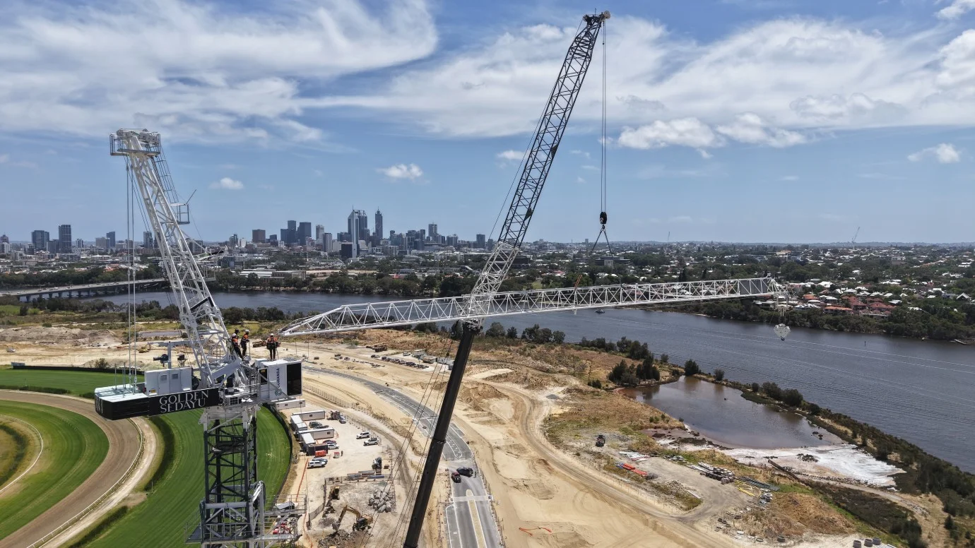 A large crane lifts a long metal structure at a construction site near a river, with a city skyline and high-rise buildings visible in the background under a partly cloudy sky.
